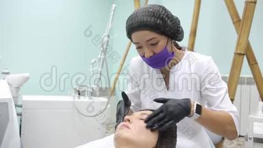 戴着刷子的粉<strong>红色</strong>手套的美容师用一个碳<strong>面膜</strong>在一个年轻女孩的脸上剥皮